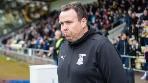 SNS Scott Kellacher is wearing a black ICT hoodie. He is standing pitchside with fans seated in a stadium behind him.