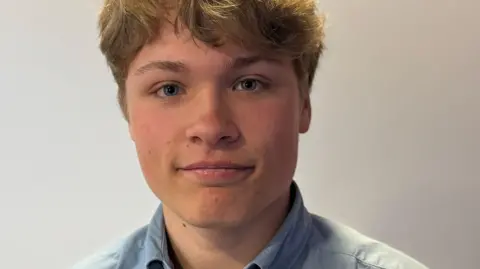Gabriel Raimondo pictured in the BBC studio. He's wearing a blue shirt and a rosette in red, blue and white. He has wavy blonde hair and is smiling. 
