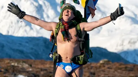 Speedo Mick Speedo Mick wearing his blue Speedos, a woolly hat, scarf, backback, gloves and nothing else, his face tilted up[wards to the sky with his eyes closed and a look of relief, against a background of snowy mountains in Glencoe, Scotland, in January 2023
