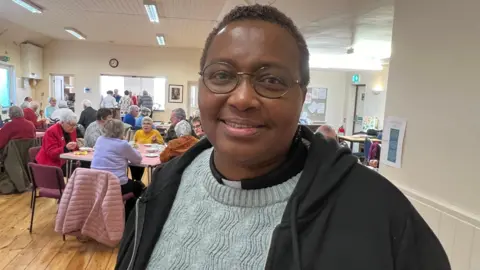 The Reverend Annie Deche. She has short dark hair and is wearing a clerical collar and a light blue jumper under a black hoodie. She is also wearing glasses. She is looking directly at the camera and smiling. 