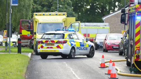 Eddie Mitchell A police car is parked in front of a house, along with ambulances and fire engines 