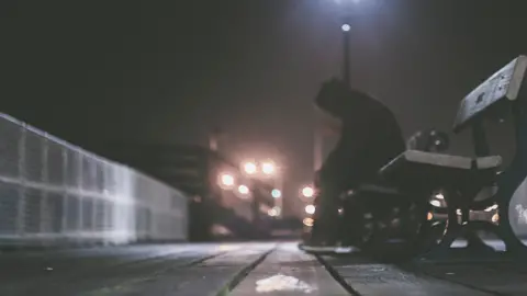 Person in silhouette sitting on a bench. Bright lights are in the background, slightly out of focus