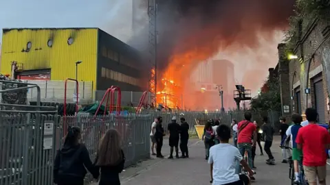 Rachael Jardine A crowd of bystanders gathers on a street to watch a large blaze sending bright orange flames and thick dark smoke into the sky behind a metal security fence, beside a yellow industrial building and brick railway arches.