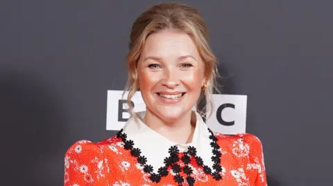 Joanna Page looking at the camera and smiling. She is wearing a red dress with a big white collar, with black flowers embroidered on it. She has blonde hair, which is tied up and gold earrings. 