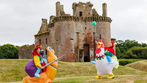 HES Two people an inflatable snail and inflatable chicken carry out a mock joust in front of Caerlaverock Castle
