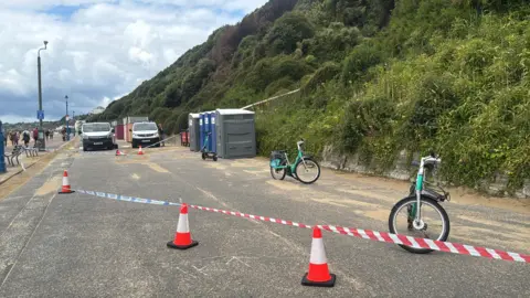 Five blue and grey portable toilets standing on a road next to a bush-covered cliff, with police tape tied to traffic cones around them, blocking the road