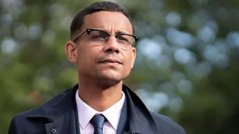 PA Media Daniel Kebede, a man in a pink shirt and blue tie with a dark blue coat and glasses. 