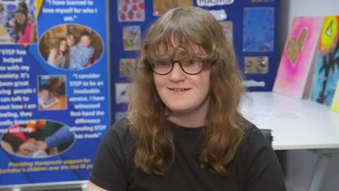 Maisie looks at the camera; She has long curly hair, fringe, glasses and wears a black t-shirt.