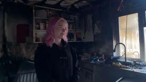 A woman in a black jacket with long pink hair inside a fire-damaged kitchen