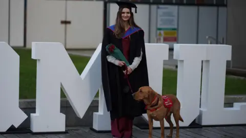 Daisy Cottle-Bailey pictured at her graduation with her medical detection dog Jarvis. Jarvis is a reddish colour and is wearing a red label identifying him as a medical detection dog. Daisy is wearing a maroon outfit and black graduation gown and mortarboard. She is stood in front of a 'QMUL' sign for Queen Mary University of London.