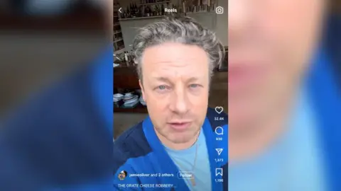 Instagram/Jamie Oliver Uma captura de tela do vídeo de Jamie Oliver no Instagram falando sobre o queijo roubado. Mostra Jamie Oliver, um homem de cabelos curtos e grisalhos, vestindo um cardigã listrado azul e uma camiseta azul