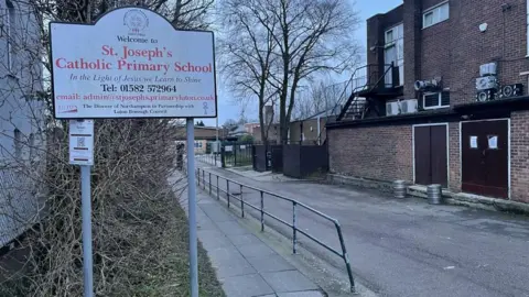 The extracurricular  of St Joseph's Catholic Primary School, showing a sign, a walkway, a ceramic  gathering  to the close    and a schoolhouse  successful  the distance. There are metallic  railings successful  the middle.