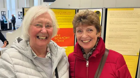 Two ladies stand in front of an information board about Gosport waterfront. One has white hair, glasses and a white coat and the other dark hair and a red coat.