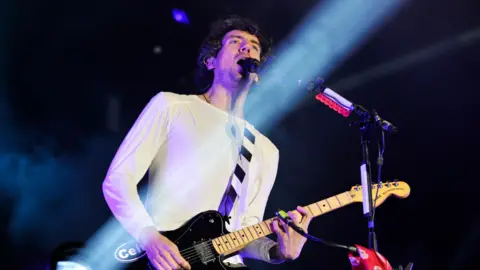 BBC Gary Lightbody of Snow Patrol plays the guitar and sings on stage. He is wearing a white long-sleeved top.