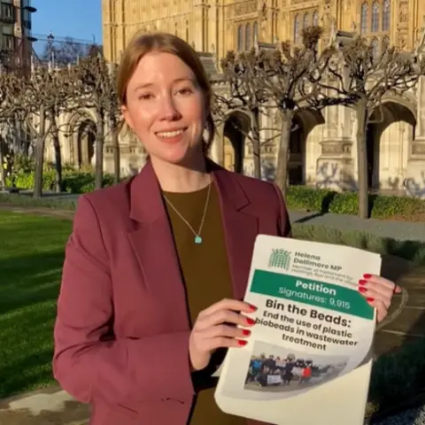 Helena Dollimore Helena Dollimore, MP for Hastings and Rye - a woman wearing a red jacket stands outside parliament carrying a poster which reads "petition, bin the beads" 