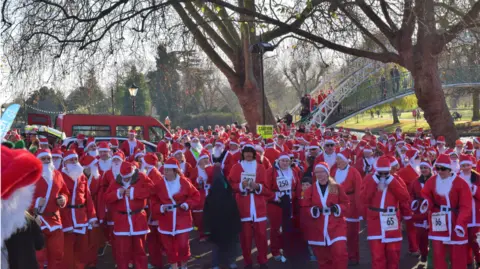 Keech Hospice Care A large group of people wearing red Santa suits with hats. All the participants have numbers on them. There is a red van and a white bridge behind them