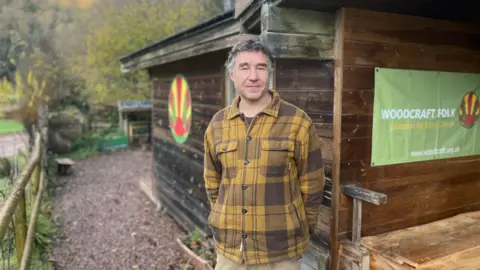 Man with brown and grey hair is wearing a mustard yellow and brown overshirt while standing in front of a wooden log cabin.