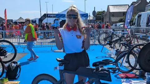Clara Isaac stands by a bike on a blue carpet. She wears dark shorts and a white t-shirt with multi-coloured sleeves. She holds up a teddy of an orca, the mascot of the games, and a bronze medal. She has long blonde hair and wears sunglasses.