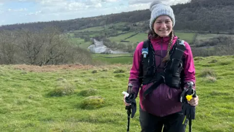 Eva Kriki Runner Eva Kriki stands wearing trail running clothing and equipment in the hills. She has long brown hair which is in plaits and wears a white woolly hat and a pink waterproof coat.