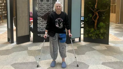 Sarah Thyer standing in what resembles a hotel lobby. She has short white hair and her glasses are resting on top of her head. She has on loose-fitting black and white patterned trousers on with a black jumper saying 'We are unlimited'. She has pale blue Croc-style shoes on and is on crutches with a blue bandage strapped to the left hand side of her hip. There are room dividers behind her, including an artwork of a jungle tree