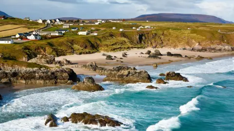 White walled properties dot an area of land near small cliffs above a beach. Waves roll in from the sea, which is a turquoise colour. There are rocky outcrops on the beach and in the sea.