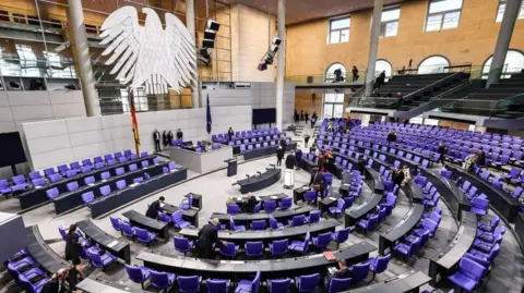 EPA Inside the Bundestag, where there are many purple chairs in a semi-circle, and a huge eagle - the logo of the Bundestag  - hanging above the speaker. There are a few people milling around but it's mainly empty.