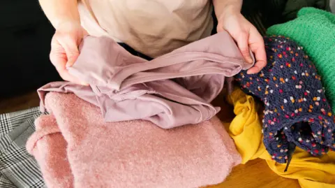 Getty Images An image of a person's torso standing in front of a pile of clothes. They are holding a light pink top, and there are pink, yellow, blue and green clothes beneath them.