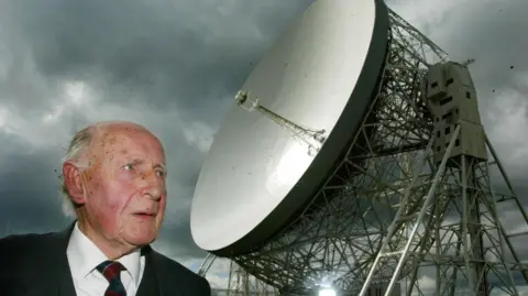Sir Bernard Lovell, with thinning white hair, stands in front of the giant Jodrell Bank telescope.