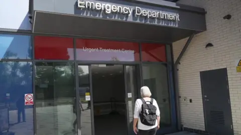 Patient walking into the Royal Bournemouth Hospital Emergency Department. The glass doors have two signs above. One says Urgent Treatment Centre, the other says Emergency Department. The person walking in is wearing a white top and is carrying a black rucksack.