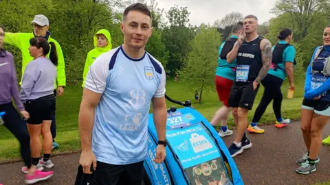 Ryan Burnett, a man with short, fair hair, poses at the start line with a weighted trolley. He is wearing a pale blue and navy t-shirt with white sleeves. The trolley has a blue cover with a photo of the late Ricky Hatton holding up a boxing belt above his head in celebration. The text on the trolley reads: "Run for Rick, 63.5 kg, Mental Health Awareness.
