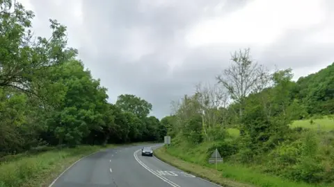 Google A three lane road swings through green countryside. A car drives towards us on one side of the road with the word "Slow" written on the road ahead of it. The other side is made of a lane with an overtaking lane in the middle with dotted white lines between them.