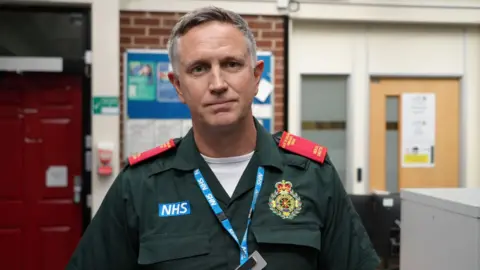 Shaun Whitmore/BBC Walsh stands in a room with a brick wall behind him. On the wall are posters and doors are either side of the wall. He is wearing green ambulance uniform with red epaulettes and the NHS logo as well as the EEAST embroidered badge on his chest.. A lanyard is around his neck. 