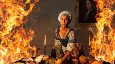 A woman looks in fear toward the camera in a scene from the play. Her clothing and the setting confirm it is set centuries in the past. On the table, there is fruit as well as two candles on display. She is bordered by flames hinting at the tone of the play. On the wall, there is a framed portrait of a man