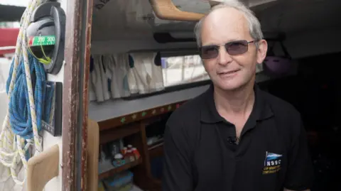 Chris sits in the cabin of the boat and looks out at the camera. There are ropes hanging on the outside. He wears dark glasses and a black t-shirt.