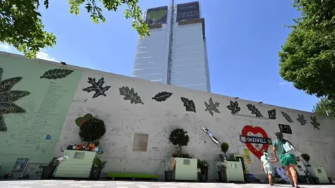 A woman and girl look up at Grenfell Tower from in front of a wall covered in written tributes