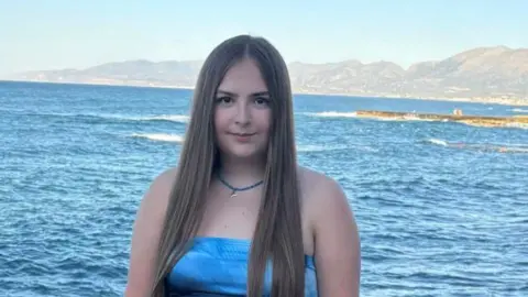 Lexie Mower Lexie Mower. She has long light brown straight hair and brown eyes. She is wearing a strapless blue top and blue necklace. Behind her is the sea and mountains part of a bay in the far distance. 