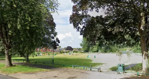A view from Cubbington Road of The Holt. There are mature trees on either side of the picture and play equipment in the centre of the image.