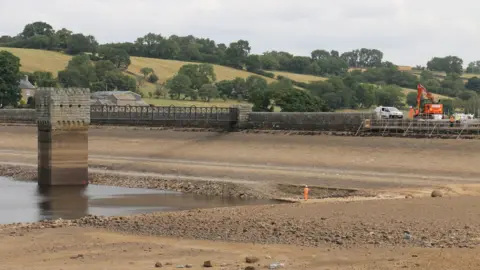 Hury reservoir. The water levels appear to be low. There is a person in orange hi-vis clothes standing at the corner - they look very small. Behind and to the right is a construction vehicle and a van next to scaffolding. 