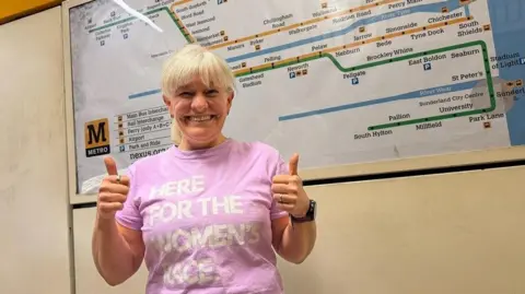 A smiling Alice Townend with blonde hair is standing in front of a large Tyne and Wear Metro map giving two thumbs up to the camera. She is wearing a pink t-shirt with the slogan 'HERE FOR THE WOMEN'S RACE' in white lettering. The setting is a Metro station with Nexus Metro branding behind.