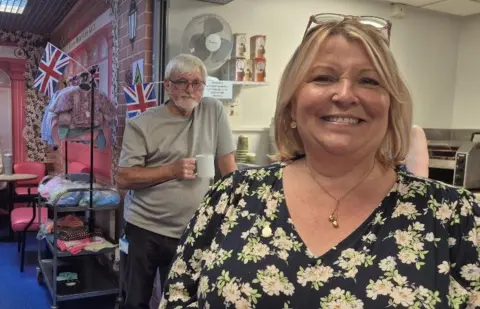 Fiona is smiling and is wearing a black dress with white flowers on it. She has short blond hair in a bob style and is standing in front of a tea and snack window, selling hot drinks and toast. There are Union flags in the background along with a man with grey hair, grey t-shirt and a cup of tea who is looking at the camera.