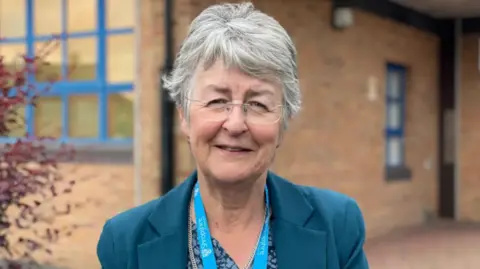 A woman with grey hair and glasses in a blue jacket in front of a brown building