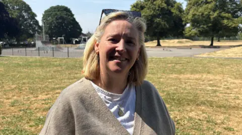 Kevin Shoesmith/BBC A woman in her early 60s with blonde hair smiles for the camerra. She is wearing a grey cardigan over a white t shirt. She is standing in a park with a skate facility behind her. 