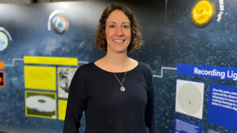 Cyrielle Opitom, a woman with short curly brown hair wearing a black boatneck top with a gold pendant necklace. She is smiling standing in front of a space backdrop.