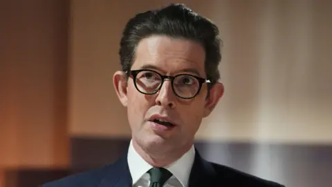 PA Media Sir Ken McCallum, a white man with dark, swept-back hair and round, dark rimmed glasses, wearing a dark suit with a white shirt and dark tie, pictured in close-up with a long lens as he delivers a speech with the background blurred behind him.