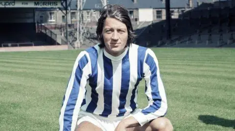 Getty Images Frank Worthington is pictured during his playing days. He is crouched on a football pitch with stands in the background. He is wearing a long-sleeved football shirt with vertical blue and white stripes, and white shorts. He is clean shaven with dark brown hair swept back.