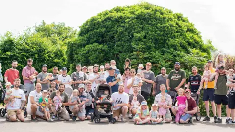 Dad's Doddle A large group of adults, children and babies pose in a group in front of a large shrub looking at the camera. Some are standing while others are kneeling or sitting. There is a dark-coloured pram in the centre of the photo.