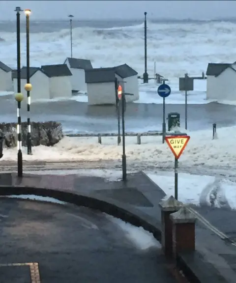 Torbay Council The photograph shows white wooden beach huts that have been moved around the seafront by powerful waves.  The huts are surrounded by sea water. In the foreground of the picture you can see where seafoam whipped up by the wind and waves meets snow that has fallen