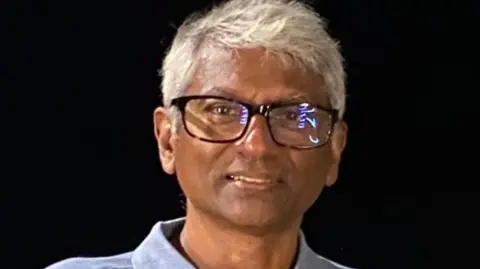 Dr Saman Perera Picture of Dr Perera wearing a blue shirt. He has white hair and is wearing glasses.