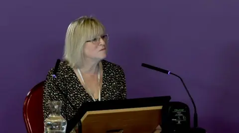 Southport Inquiry Joanne Hodson, who has shoulder length blonde hair, a fringe and glasses, sits at a witness table in front of a screen and a black microphone in front of a purple screen. 