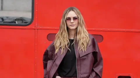 PA Media Cara Delevingne arrives at the Topshop catwalk show. She is wearing a purple leather jacket and posing in front of a red London bus.